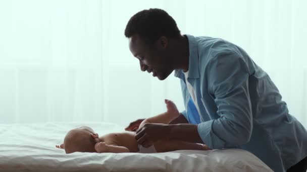 Счастливый черный отец меняет подгузник для своего новорожденного ребенка и забирает его из постели, проводит время дома, замедленная съемка — стоковое видео