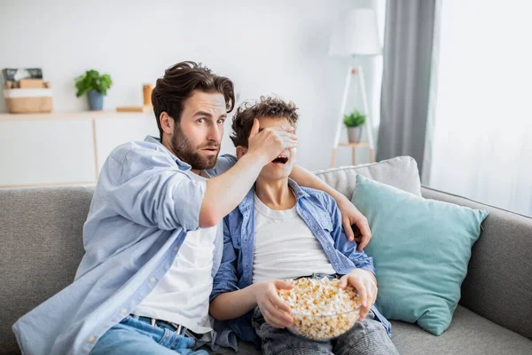 Father covering his sons eyes from inappropriate content while watching movie on tv with popcorn