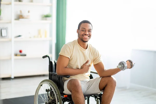 Physical rehabilitation for disabled people. Handicapped black guy in wheelchair making exerises with dumbbell at home