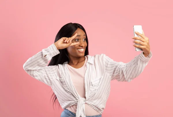 Joyful black woman taking selfie with smartphone and showing victory gesture on pink studio background