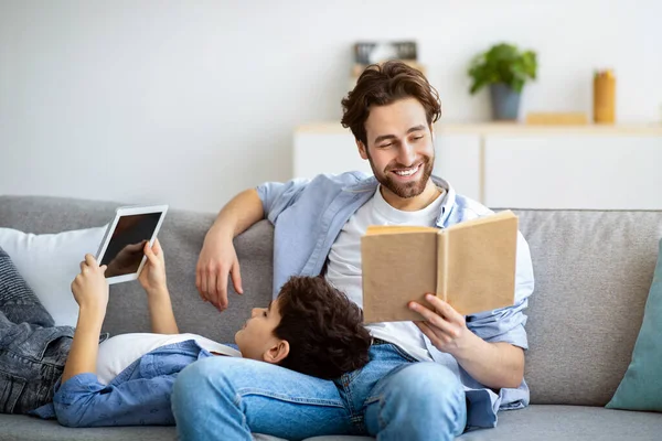 Different generations. Father reading book while his son using digital tablet, sitting together on sofa