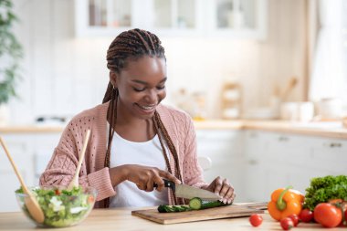 Mutfakta taze sebze salatası pişiren, salatalık doğrayan mutlu genç zenci kadın.