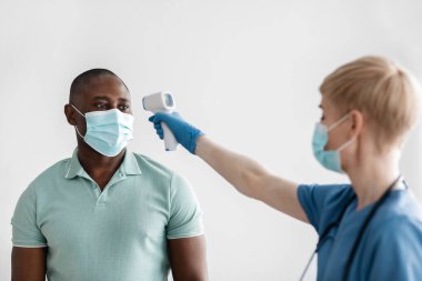 Koruyucu maskeli, eldivenli ve üniformalı orta yaşlı kadın sıcaklığı modern kızılötesi termometreyle ölçüyor.