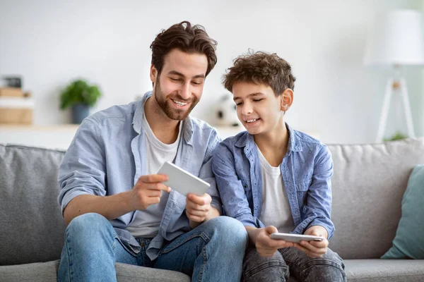 Show me your score. Boy competing with dad in online video games on smartphones, sitting on couch at home