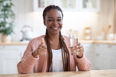 Neşeli siyah kadın mutfaktaki bardaktan su içiyor ve başparmağını kaldırıyor.