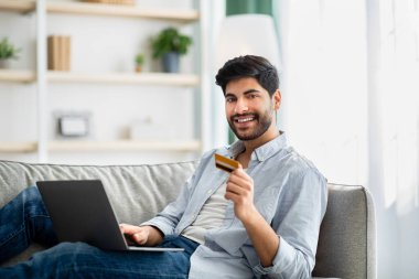 Çevrimiçi ödeme. Gülümseyen Arap adam dizüstü bilgisayar ve kredi kartı kullanıyor, eve para aktarıyor.