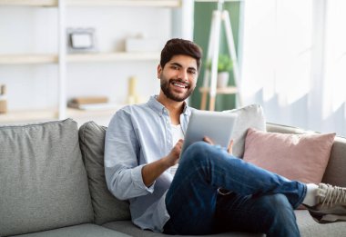 Boş zaman kavramı. Arap adam dijital tablet kullanıyor, oturma odasındaki rahat koltukta dinleniyor.