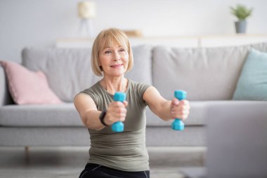 Evde dizüstü bilgisayarın yanında halterli, spor videolarına online fitness yapan olgun kadın eğitimi.