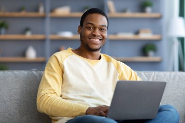 Evdeki kanepede dizüstü bilgisayarı olan neşeli siyahi adam.