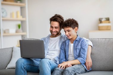 Mutlu baba ve oğul dizüstü bilgisayar kullanıyor, izlemek için film arıyor, internette geziniyor ve evde birlikte vakit geçiriyorlar.