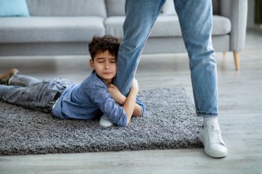 Gitme, baba. Üzgün çocuk babasının bacağını kucaklıyor, babasının yanında kalması için yalvarıyor, halının üzerinde yatıyor.