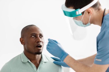 Üniformalı genç bir doktor, koruyucu eldivenler ve maske takıyor. Afrikalı Amerikalı bir hastadan test yaptırıyor.