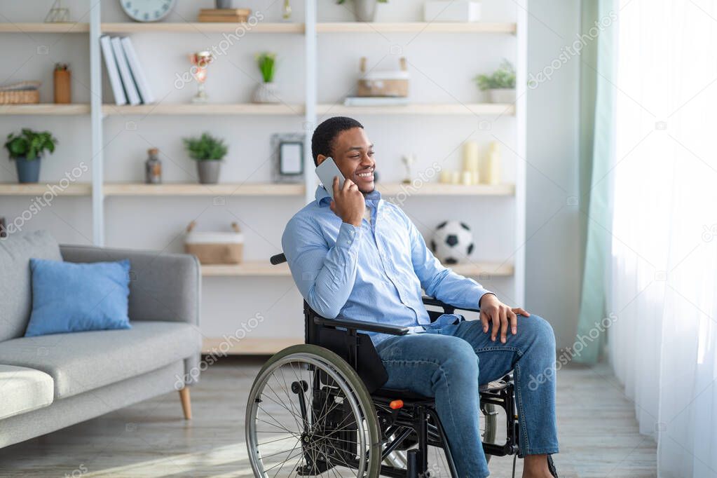 Joyful black guy in wheelchair having phone conversation with friend at home