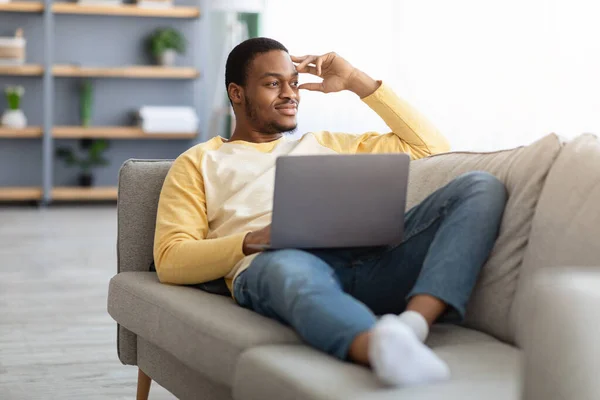 Dreamy black guy with laptop resting on sofa at home