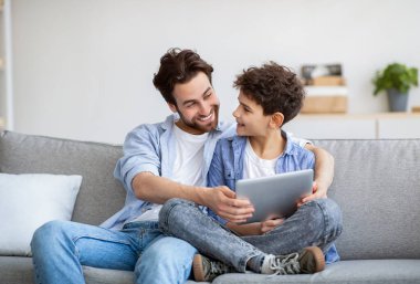 Aile zamanı. Mutlu baba oğluna tablet bilgisayarda özel uygulama kullanmayı öğretiyor. Kanepede yan yana oturuyor.