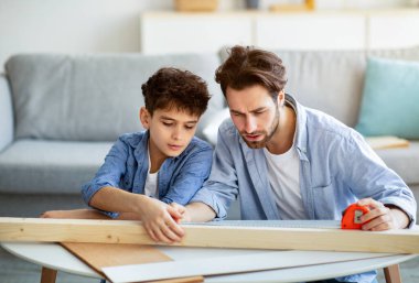 Yoğunlaşmış baba ve oğul tahta parçasını bantla ölçüyor, çocuk ev işi öğretiyor.