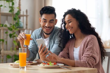 Orta Doğulu çift mutfakta kahvaltı ederken akıllı telefondan fotoğraf izliyorlar.