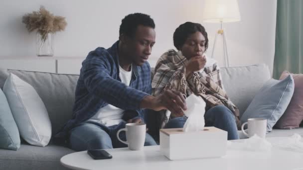 Young african american woman feeling sick, coughing on couch, caring husband supporting her and giving tissue — Vídeo de stock