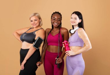 Farklı vücutlara sahip çok ırklı kadınlar bej arkaplan üzerinde spor kıyafetleriyle poz veriyorlar.