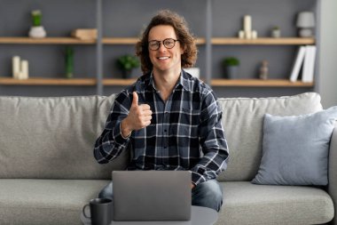 Dizüstü bilgisayardaki serbest çalışan mutlu adam evde otururken başparmak hareketi yapıyor.