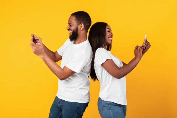 African american couple using smartphones, yellow studio wall