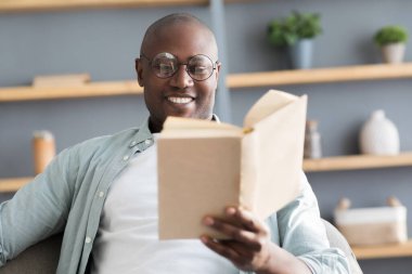 Boş zaman konsepti. Siyahi olgun adam kitap okuyor, uzun kurgu romanlarının tadını çıkarıyor, en çok satanlar listesi, seçici odaklanma.