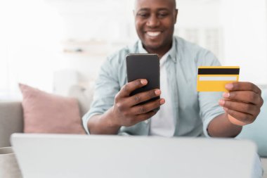 Banka işlemleri. Mutlu Afrikalı Amerikalı adam kredi kartı tutuyor ve akıllı telefonu evde alışveriş için kullanıyor.