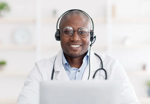 Online medical blog from home. Happy black doctor in headphones, sitting at table in interior of living room