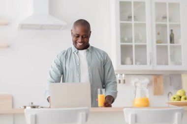 Mutlu Afro-Amerikalı olgun bir adam portakal suyu içiyor ve dizüstü bilgisayarla mutfakta film izliyor.