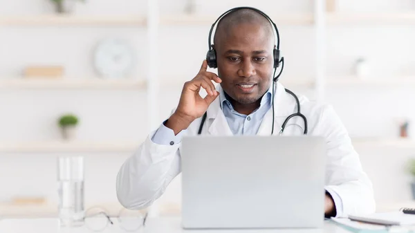 Hotline in hospital. Smiling black male doctor in white coat, wearing headphones and looking at laptop computer
