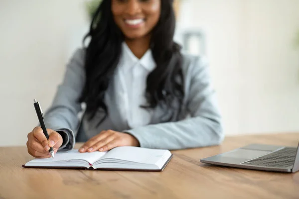 Unrecognizable black female secretary writing down info during online business meeting at workplace, selective focus