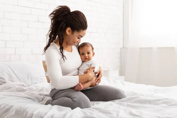 Black Mother Embracing Adorable Baby Toddler Sitting On Bed Indoor