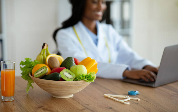 Nutritionist working on laptop, giving online consultation to client at clinic, focus on bowl of fruits and vegetables