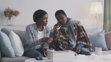 Genç Afro-Amerikalı adam termometreyle ateşi ölçüyor, ilgili eş endişeli ve rahatsız hissediyor.