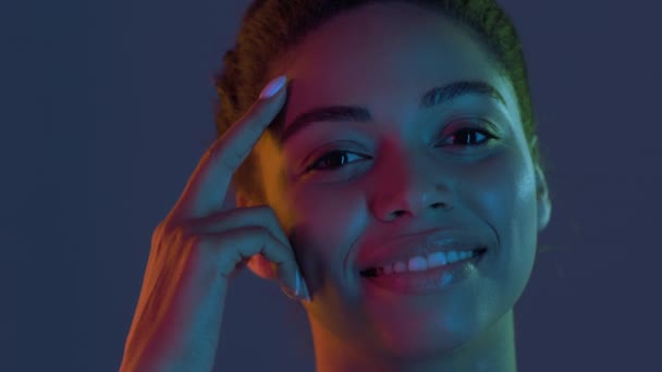 Skönhetsvård. Charmig ung afrikansk amerikansk dam smeker hennes släta ansikte och ler mot kameran i neonljus — Stockvideo