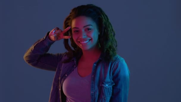 Nocne życie. Szczęśliwa młoda Afroamerykanka tańcząca, gestykulująca znakiem pokoju, stojąca w neonach — Wideo stockowe