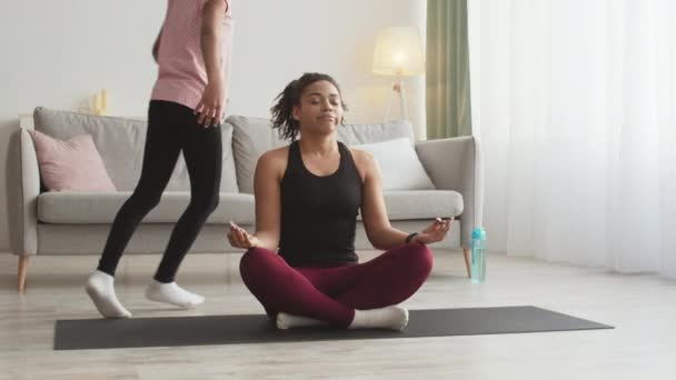 Jovem afro-americana mãe meditando, sentado no chão em casa, filha ativa correndo ao redor e perturbando-a — Vídeo de Stock