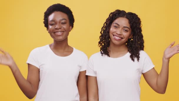Duas amigas afro-americanas sociáveis acenando as mãos para a câmera e sorrindo, gesticulando Olá, fundo amarelo — Vídeo de Stock