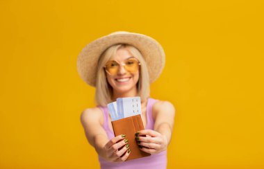 Elinde pasaport ve sarı stüdyo arka planına karşı uçuş biletleri olan aşırı neşeli bir bayan yolcu.