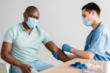 Koruyucu maskeli milenyum doktoru yetişkin Afrikalı Amerikalı hastaya iğne yapıyor.