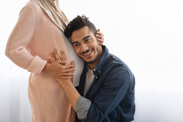 Happy eastern man embracing tummy of his pregnant wife, listening baby kicks