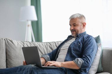 Kanepede dinlenirken dizüstü bilgisayar kullanan neşeli, olgun bir adam.