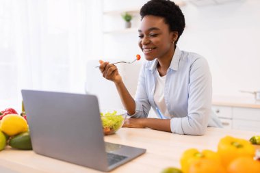 Modern mutfakta dizüstü bilgisayar kullanan, salata yiyen mutlu siyah kadın.