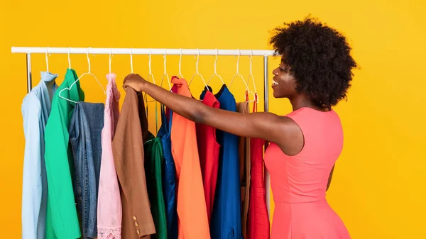 African Woman Shopping Choosing Clothes On Clothing Rail, Yellow Background