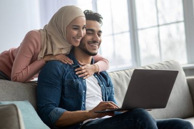Başörtülü kadın yakışıklı kocasına dizüstü bilgisayarla sarılıyor.