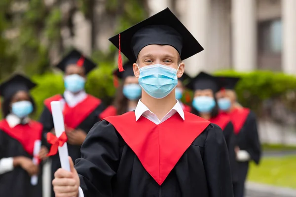 Guy student in graduation dress showing diploma, wearing face mask