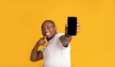 Neşeli orta yaşlı Afro-Amerikalı beyaz tişörtlü yakışıklı erkek, parmaklarını telefonda gösteriyor.