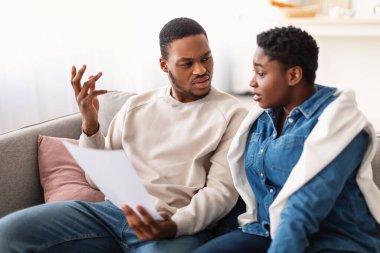 Kafası karışmış afro bir çift evde kitap okuyor, kanepede oturuyor.