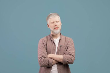 Kollarını kavuşturup poz veren genç albino adam, turkuaz arka planda kameraya ciddi bir yüz ifadesiyle bakıyor.