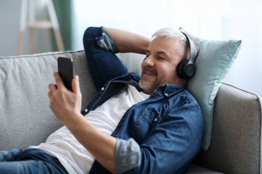 Rahatlamış, gri saçlı, kulaklıklı adam akıllı telefondan müzik dinliyor.